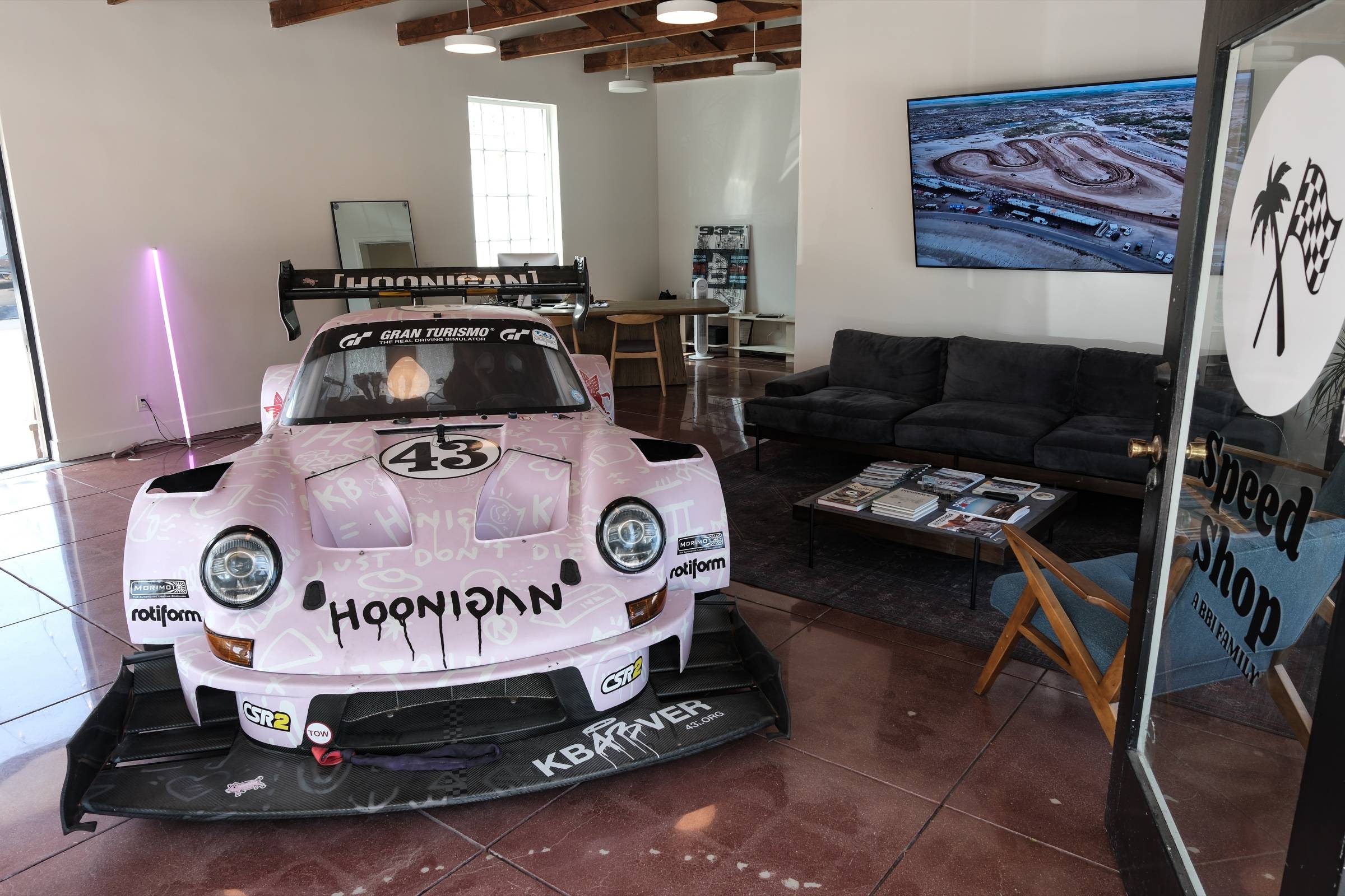 The HB Speed Shop lounge: a pink Hoonigan Porsche 911 parked on the polished floor beside a sofa, the shop logo on the glass door.