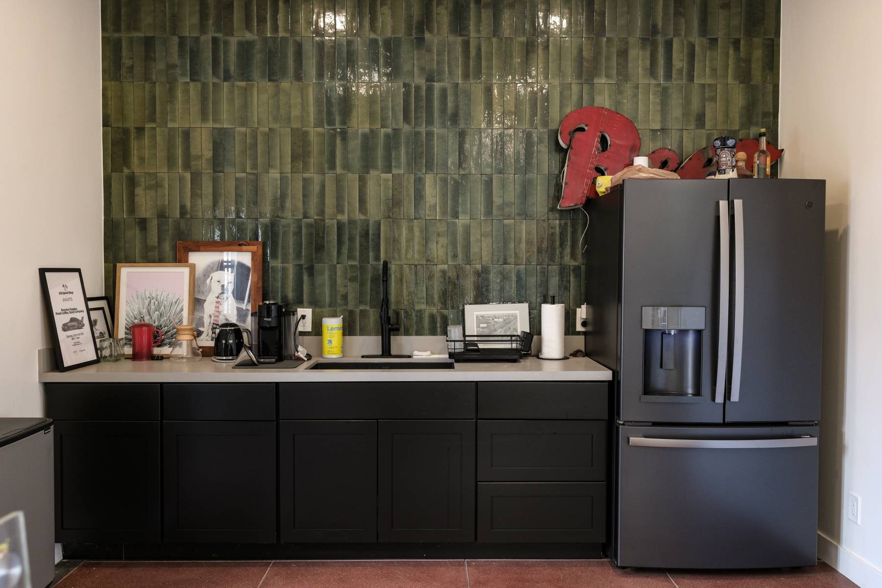A kitchenette with dark cabinets, a sink, and a fridge against a green tiled wall.