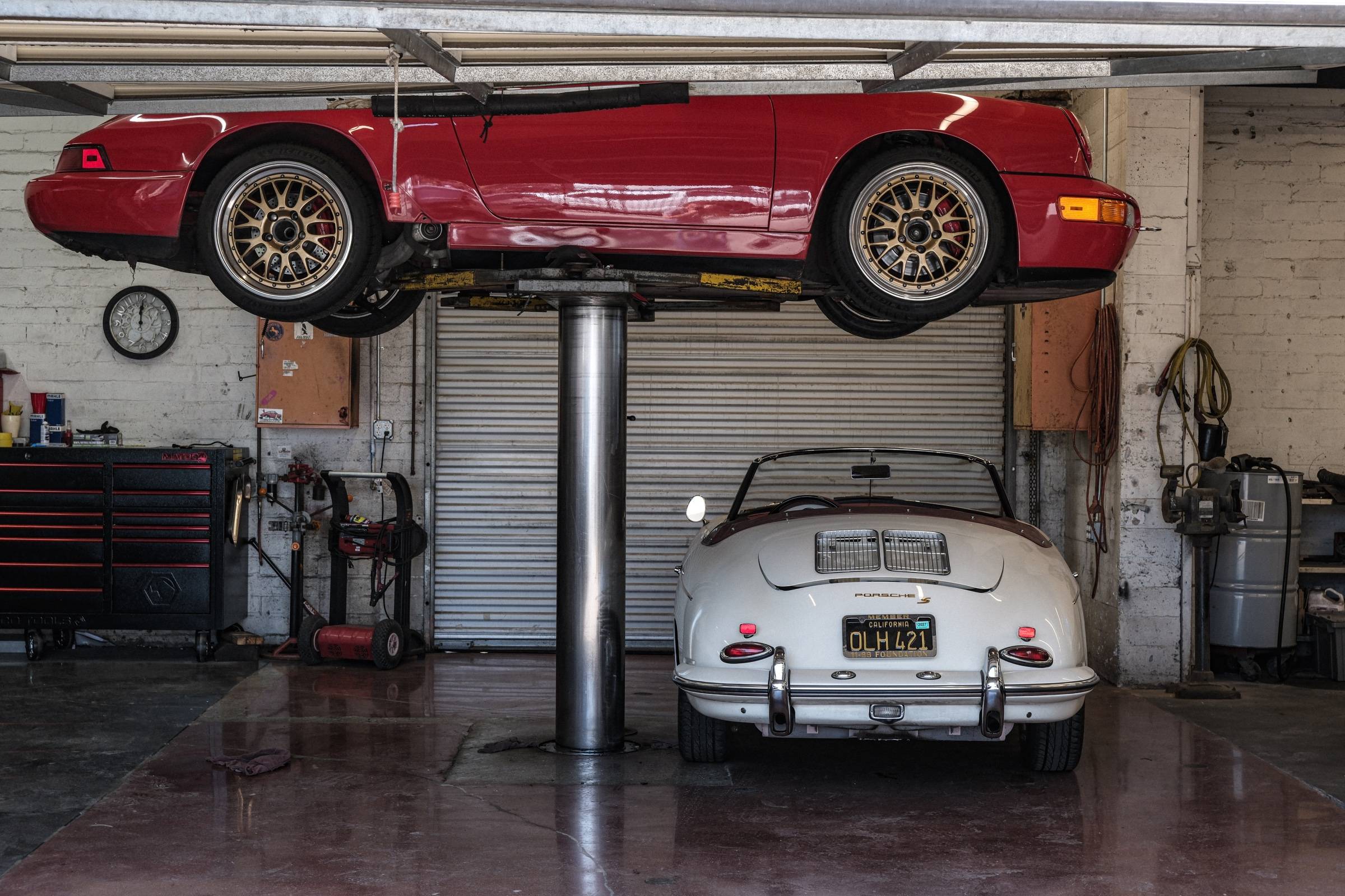 Inside the shop: a red Porsche 911 raised on the lift above a white Porsche 356, toolboxes along the wall.