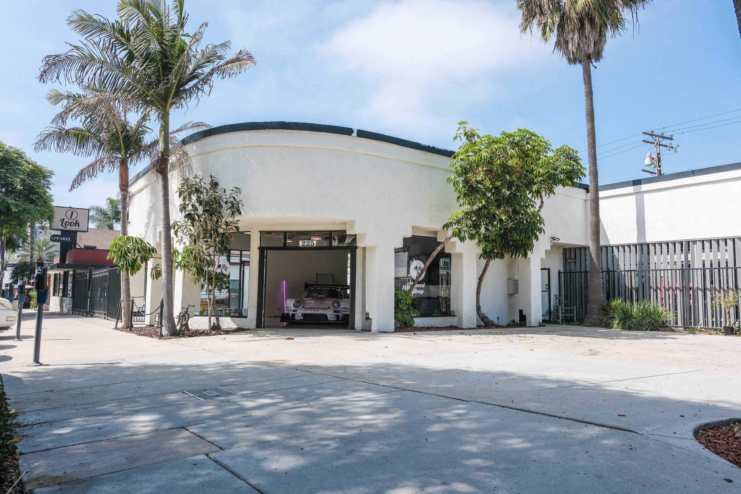 The HB Speed Shop building at 225 5th St — white curved storefront under palm trees, roll-up door open with a Porsche 911 inside.